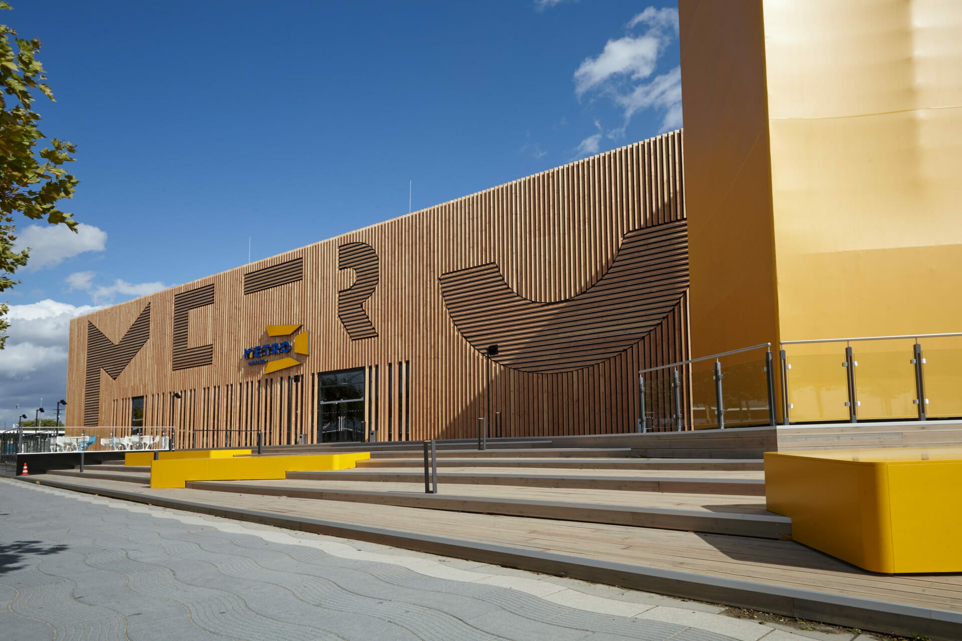 Exterior view of a modern, yellow building with wooden accents and signage that reads "MEET" and "Open365." The METRONOM-inspired building is surrounded by a paved sidewalk and a staircase leading up to the entrance.