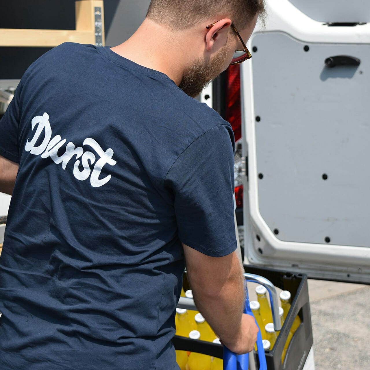 A man in a blue "Durst" t-shirt loads a box with water bottles into the back of a white van, preparing for another busy day at the marketplace.