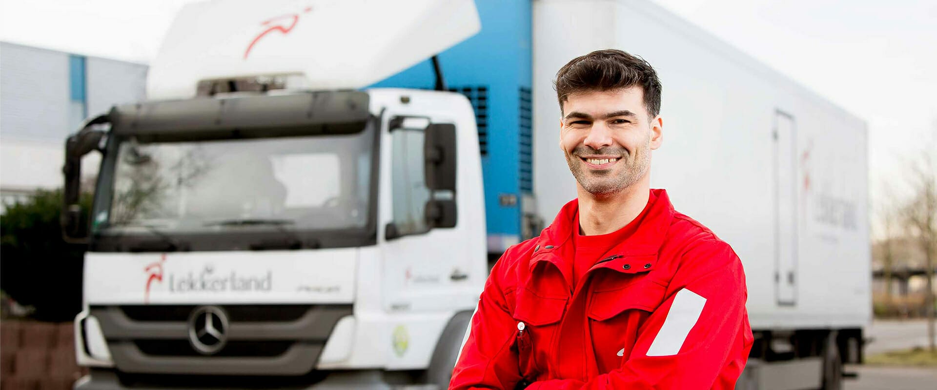 A man in a red jacket folds his arms and smiles in front of a white Lekkerland delivery truck with a logo on the side.