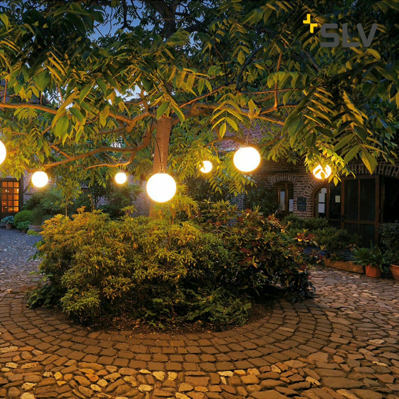 A courtyard at dusk with a large tree adorned with glowing spherical lights hanging from its branches, surrounded by cobblestone pathways and dense foliage, evokes the serene ambiance of a digital company’s tranquil retreat.