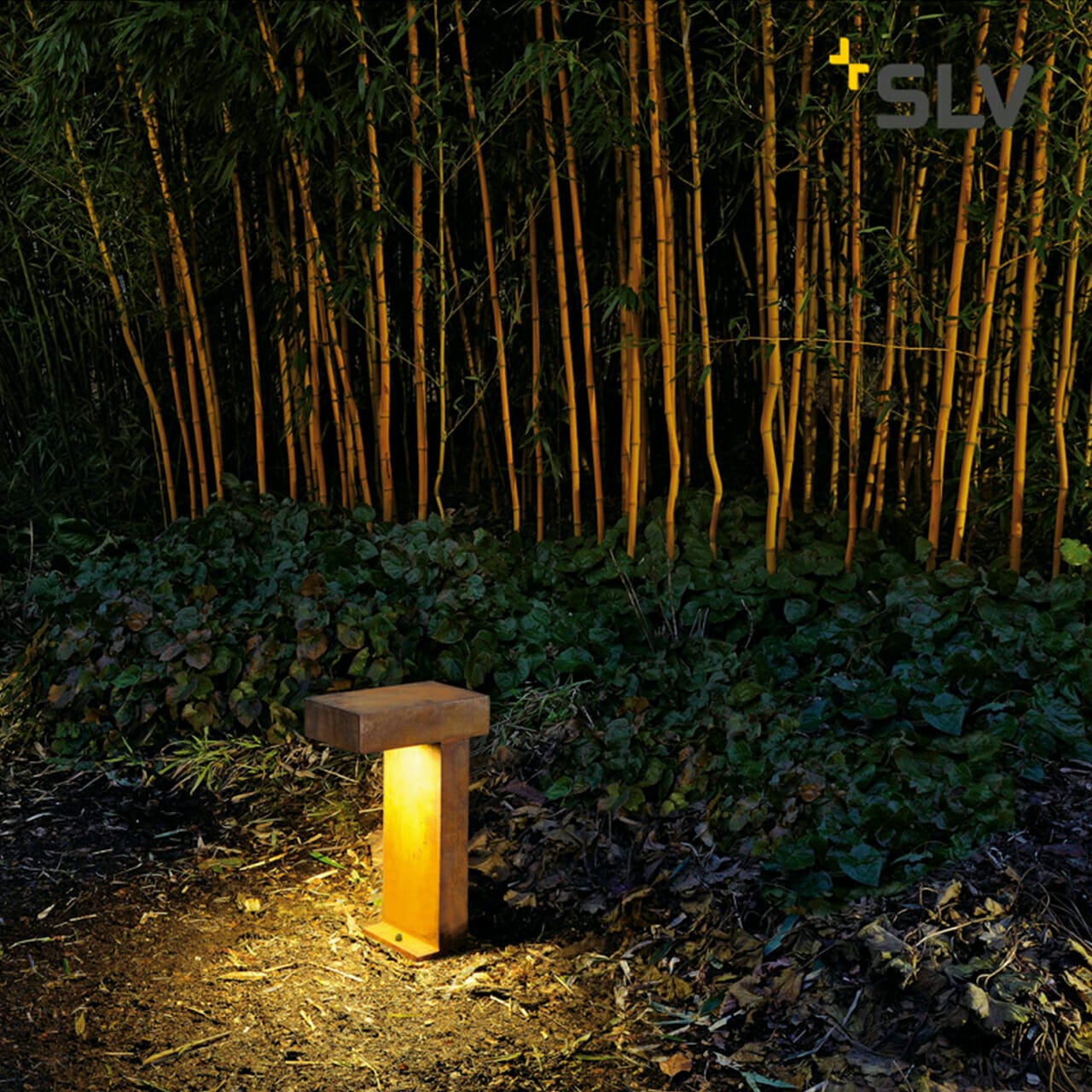 A wooden outdoor light fixture illuminates the ground near dense bamboo plants at night, showcasing a serene ambiance that merges nature with technology. The SLV logo of this innovative digital company is visible in the top right corner.