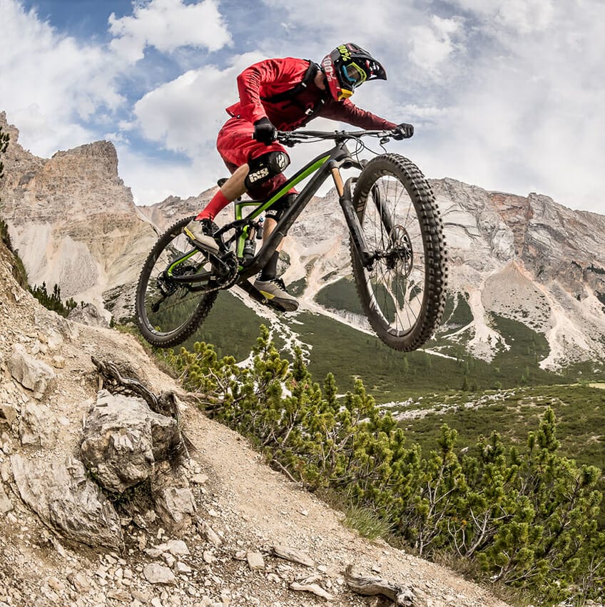 A person in red gear and a helmet, riding a state-of-the-art ROSE Bike, is mid-air on a mountain bike, performing a jump on a rocky trail with mountainous terrain and a cloudy sky in the background.