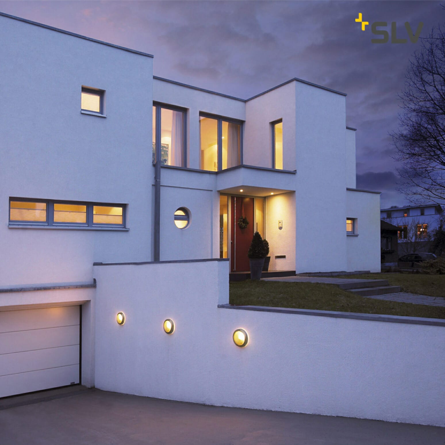 Modern two-story white house at dusk with illuminated exterior lights and a garage on the left. The building, reminiscent of high-end designs often featured in mvp cases, has large windows and a flat roof design. The sky is partly cloudy.