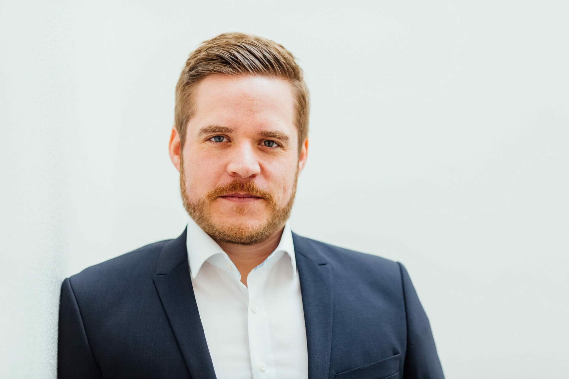 A man with short hair and a beard in a white shirt and dark blazer looks at the camera against a plain background, ready to present at the K5 Konferenz 2022.