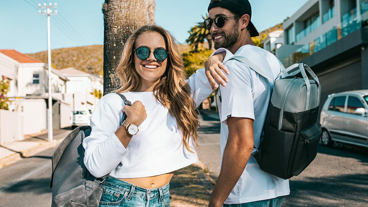 Two people wearing Kapten & Son sunglasses and backpacks smile while standing on a sunny street in a residential area.