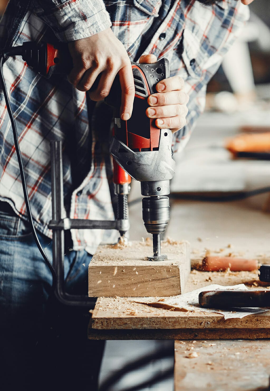 A person wearing a plaid shirt engages in some DIY, using a power drill on a piece of wood in a workshop. Various tools and materials are scattered on the workbench.