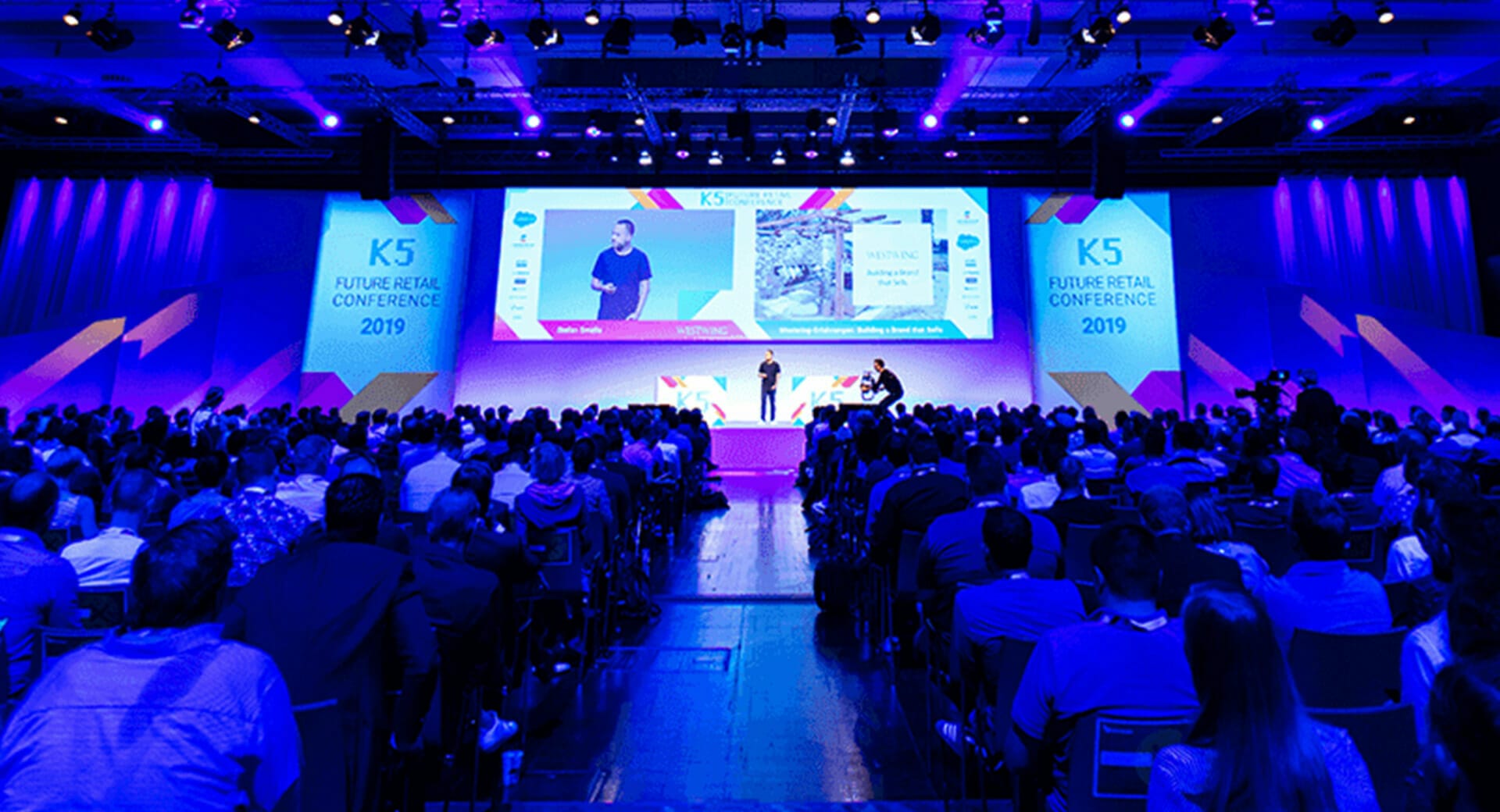 A speaker stands on stage giving a presentation at the K5 Konferenz 2023, facing a large audience seated in a dimly lit conference hall.