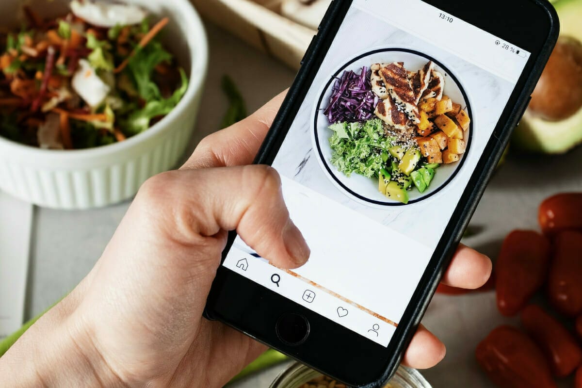 Close-up of a hand holding a smartphone displaying a photo of a healthy meal with grilled chicken, quinoa, and assorted vegetables. In the background, there are bowls of salad and various vegetables, illustrating convenient online grocery shopping insights for fresh ingredients.