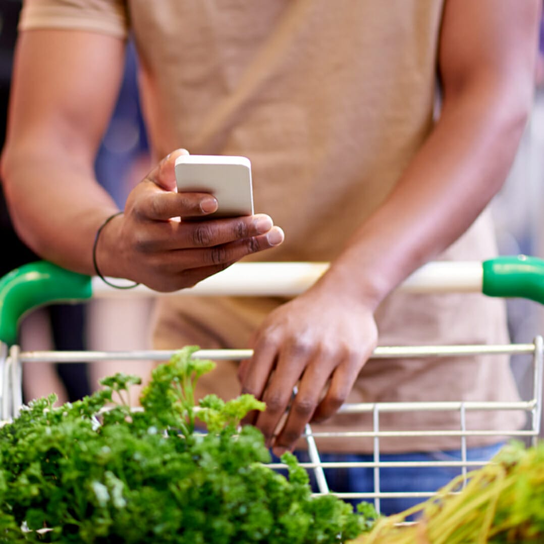 A person is navigating the aisles of METRO, pushing a shopping cart filled with green vegetables while using a smartphone with their other hand.