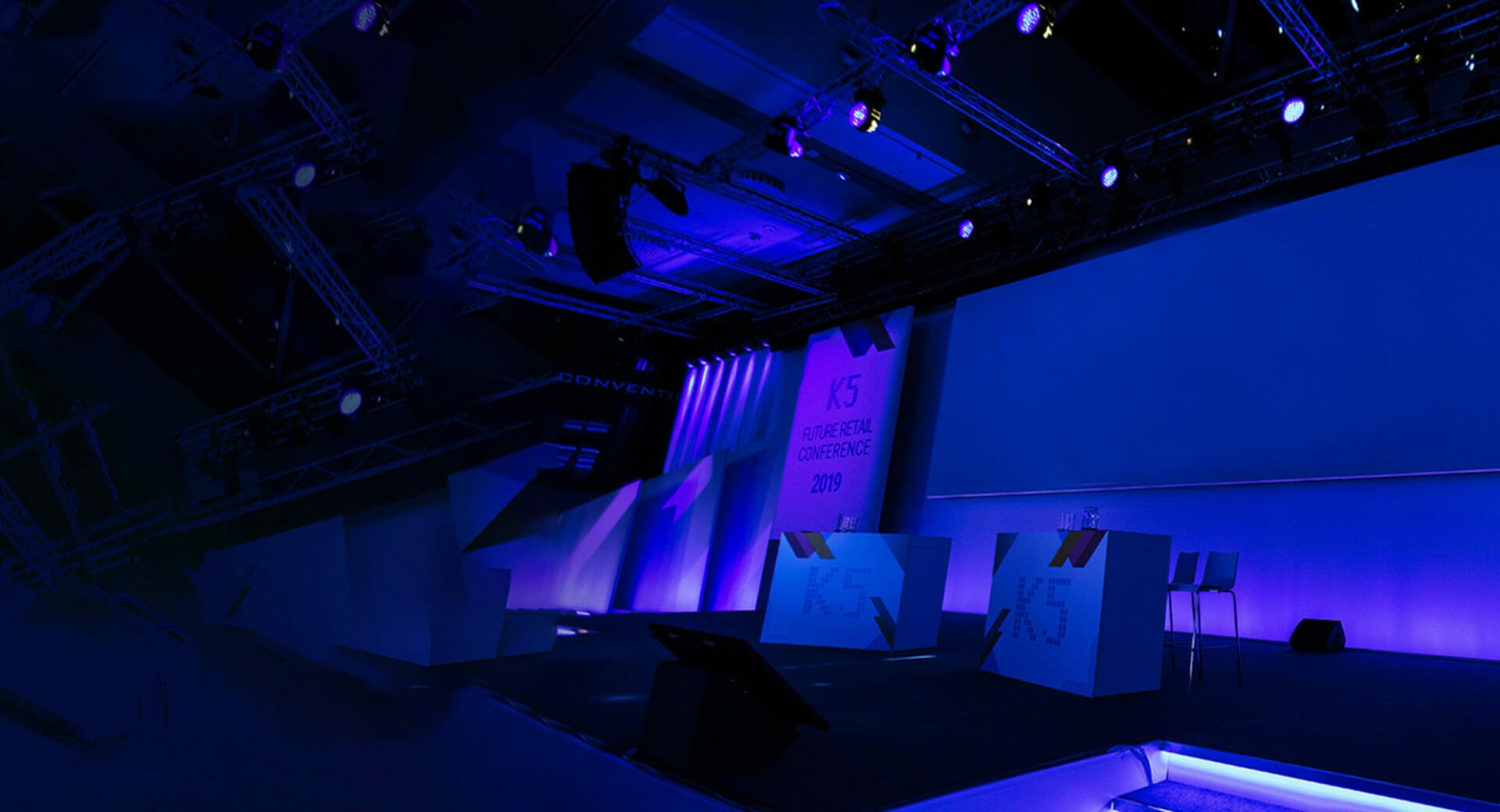 A stage setup with purple lighting for the K5 Conference 2019 in Saudi Arabia. Two podiums and a few barstools are seamlessly arranged on the stage.