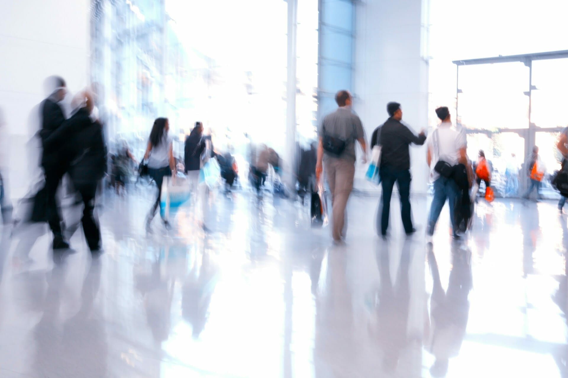 People walking through a bright, modern corridor with large windows, some carrying Intersport bags. The shiny floor reflects their movements.