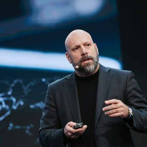 A man with a beard wearing a dark suit speaks on stage, holding a presentation remote. He has a microphone attached to his ear. The background is blurred and includes a large display screen showcasing details about the Partner Program.