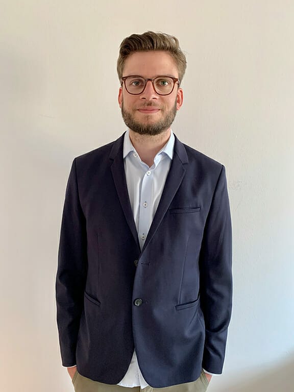 A man with glasses and a beard is wearing a dark blue blazer over a white shirt. He stands against a plain light-colored background, exuding a DIY sense of style.