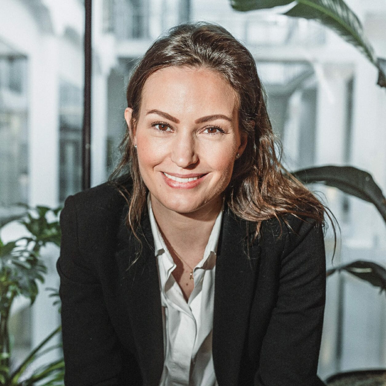 A woman in a black blazer and white shirt smiles while sitting indoors with leafy plants in the background, possibly reflecting on her recent experience at the OMR Festival.