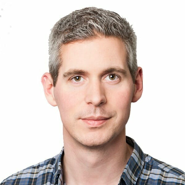 A man with short gray hair and a slight smile, wearing a blue and gray plaid shirt, is looking directly at the camera against a plain white background. His approachable demeanor EXCITEs confidence and trust.