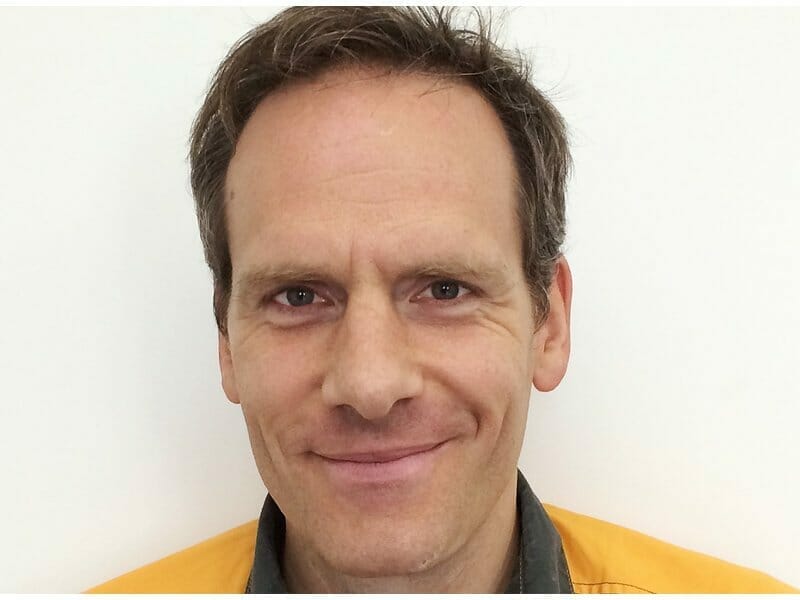 A man with short brown hair, wearing a yellow shirt, smiles while looking at the camera against a white background, ready for his onair appearance.