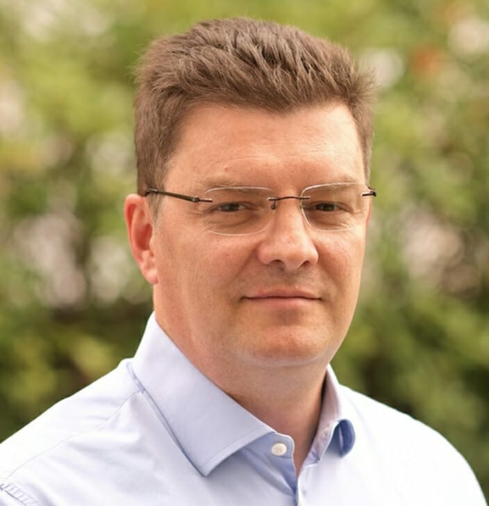 A man with glasses and short, brown hair is wearing a light blue shirt, exuding a Spryker vibe. The background is blurred foliage.