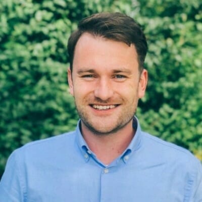 A man with short brown hair, wearing a light blue button-up shirt, smiles excitedly in front of a green, leafy background.