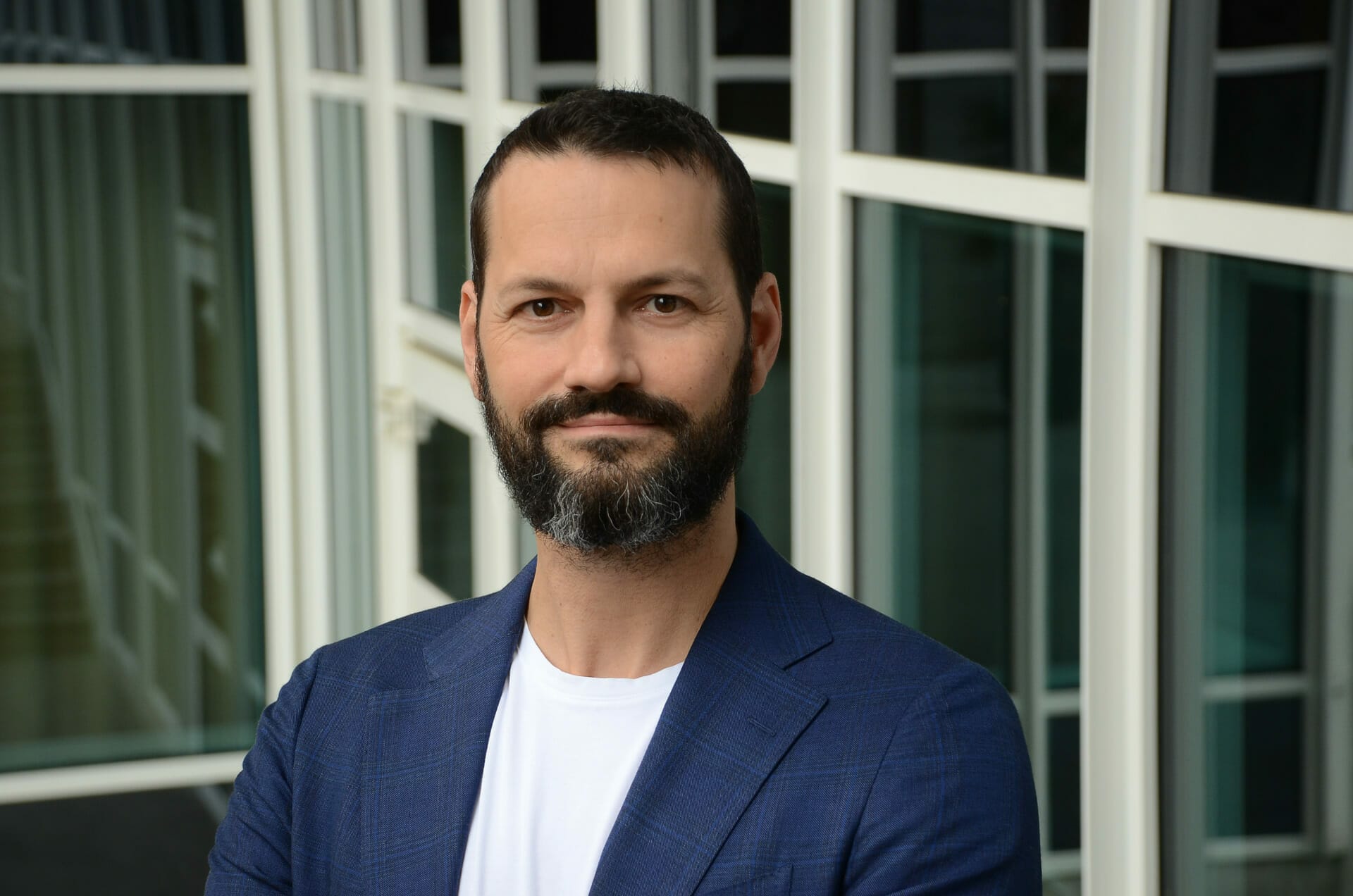 A man with a beard and short hair is wearing a blue blazer and white shirt, standing in front of a building with large windows, exuding an air of quiet confidence as though he's ready to EXCITE the world with innovative ideas from Spryker.