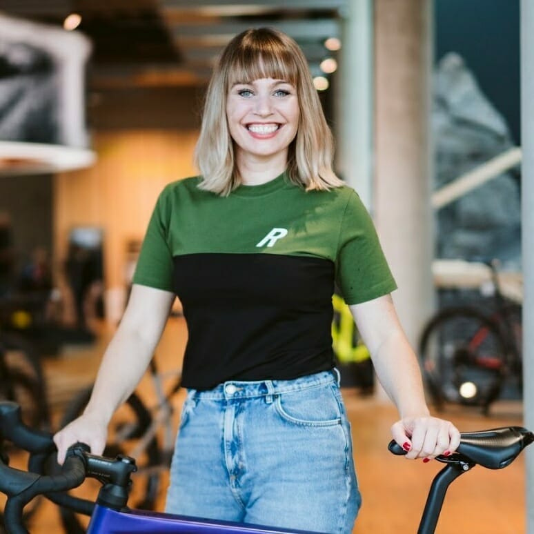 A smiling person with shoulder-length blonde hair stands indoors, holding a bicycle handlebar. They are wearing a green and black t-shirt with a letter "R" on it and blue jeans, looking ready to EXCITE those around them.