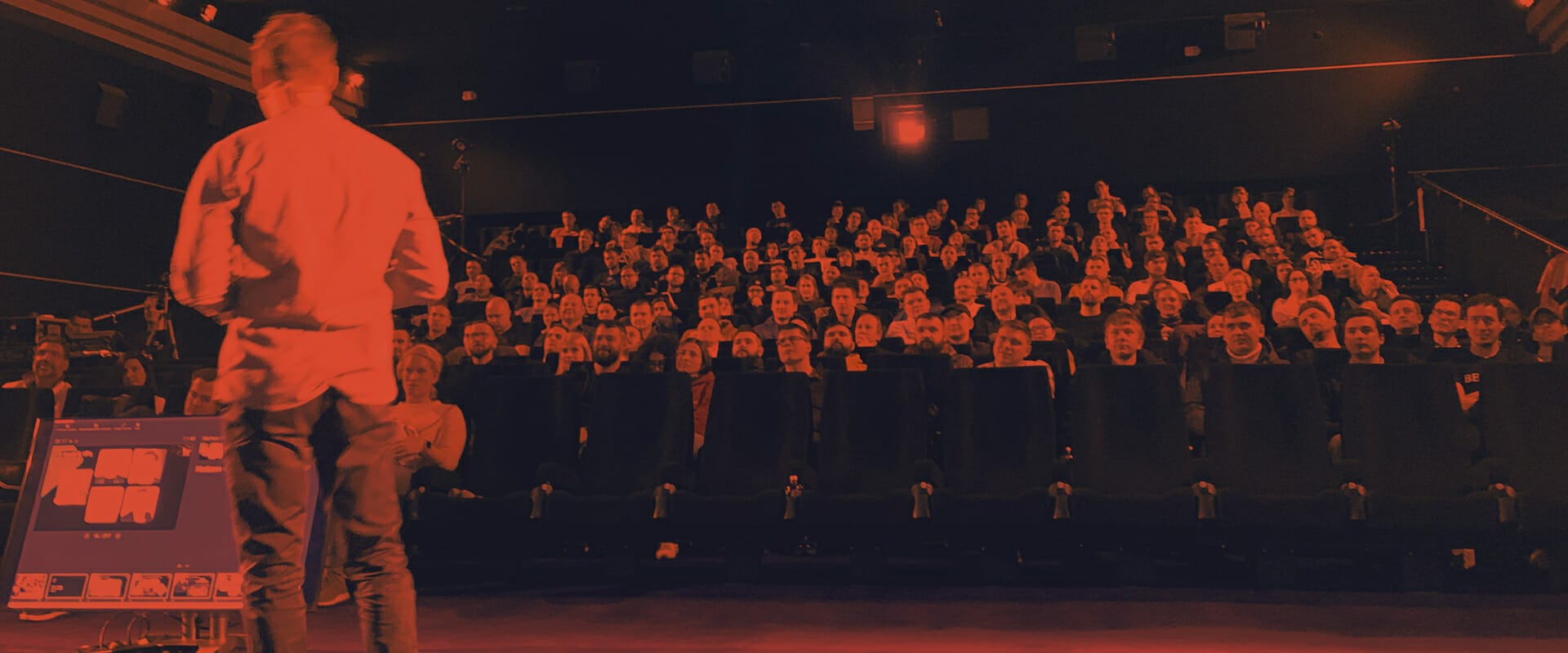 A person stands on stage at a Spryker event, addressing an audience seated in an auditorium. The audience is facing forward, focused on the speaker with excitement. The image is tinted with a red hue.