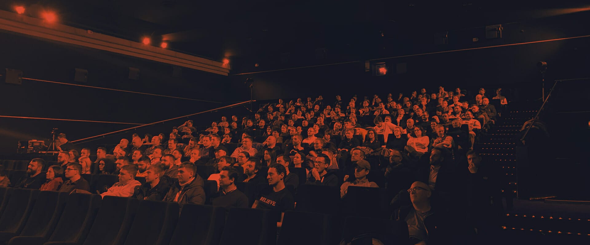 A crowd of people, EXCITE filling the dimly lit auditorium, attentively facing the front.