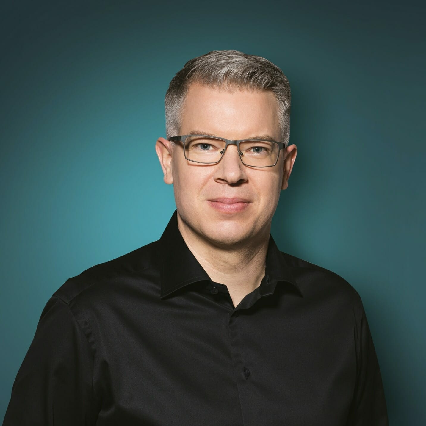 A man with short gray hair and glasses, wearing a black Spryker shirt, stands against a solid teal background, ready to EXCITE any event.