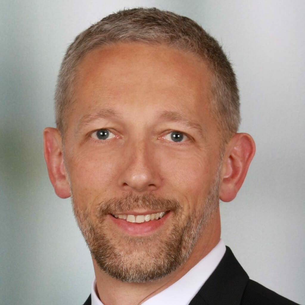 A man with short gray hair and a beard, wearing a dark suit jacket, white shirt, and patterned tie, smiles at the camera against a light background, embodying the essence of EXCITE.