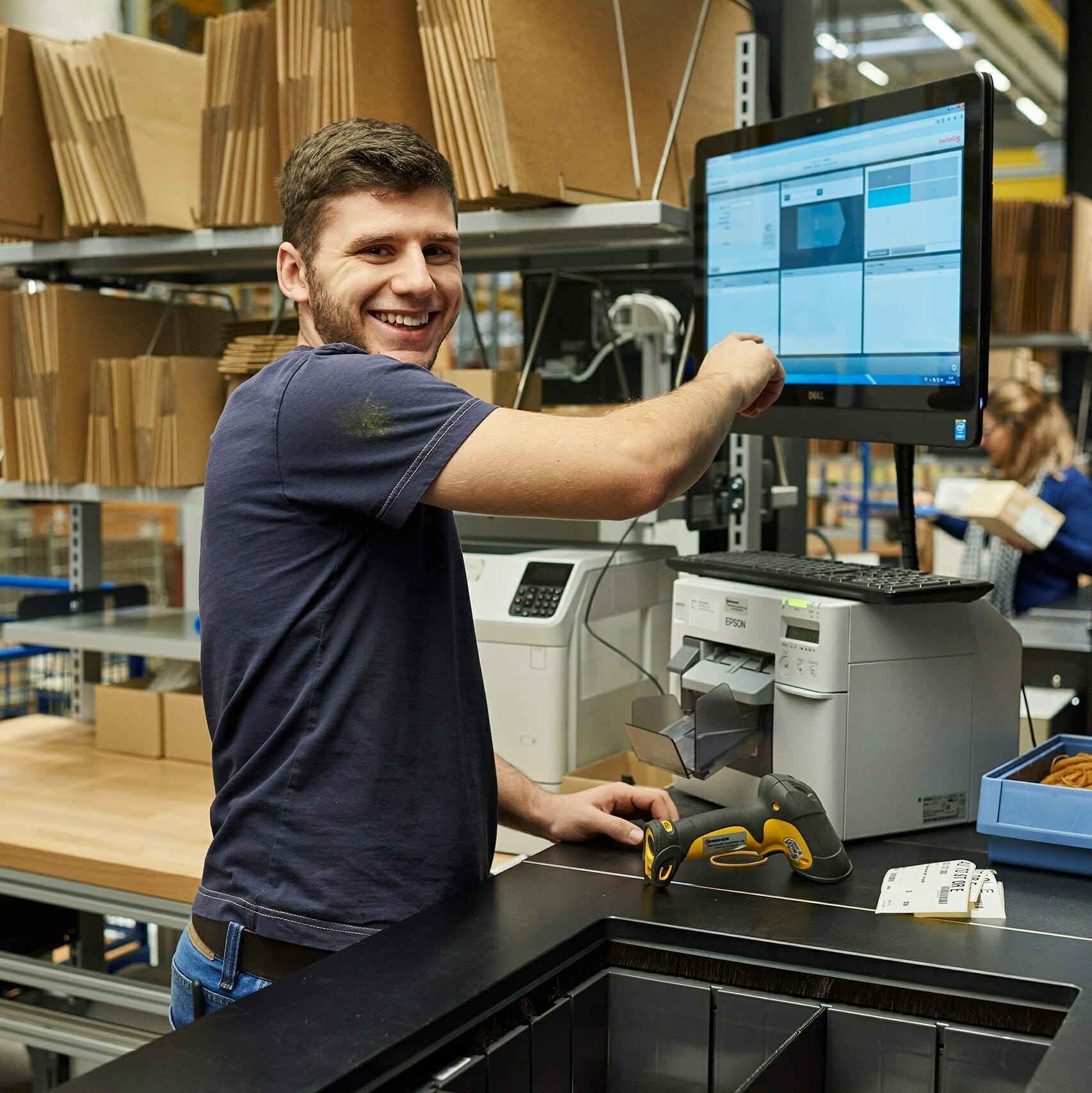 A person stands in a warehouse, smiling and pointing to a computer screen displaying various images. A barcode scanner and a label printer are on the desk beside them. Shelves with boxes are in the background, marked clearly by Winterhalter+Fenner labels.