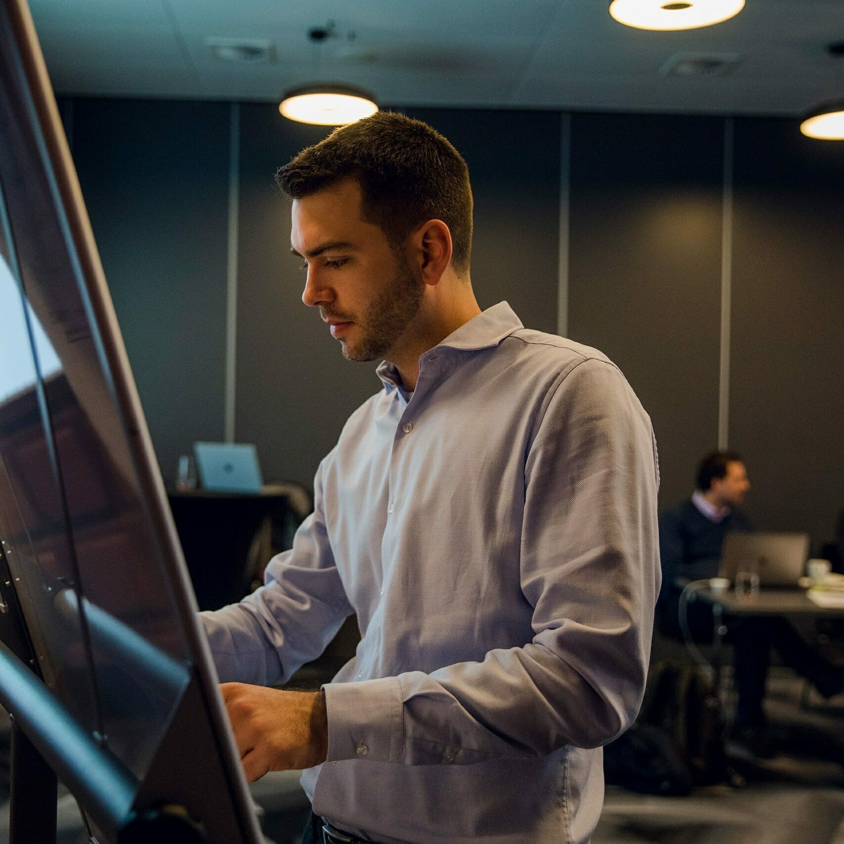 A man in a light blue shirt works intently on a large touchscreen display in a modern office setting, possibly analyzing Winterhalter+Fenner data. In the background, another person is seated, focused on their laptop.