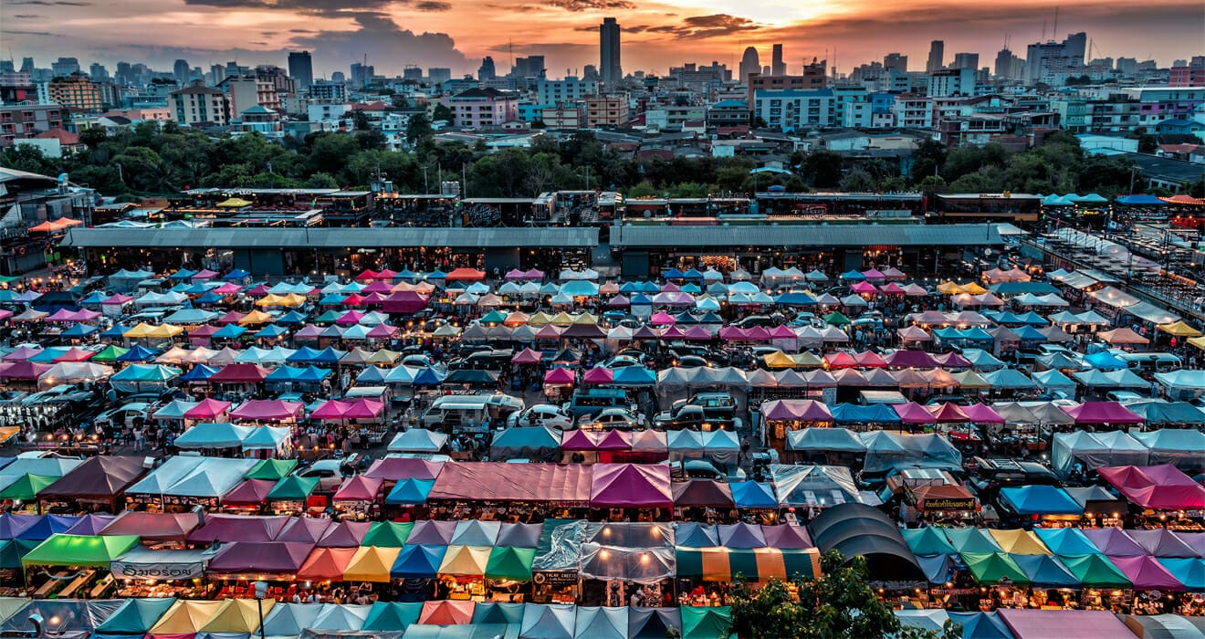 Aerial view of a crowded outdoor market with colorful tents, set against a city skyline at sunset, epitomizing the Universal Marketplace Solution.