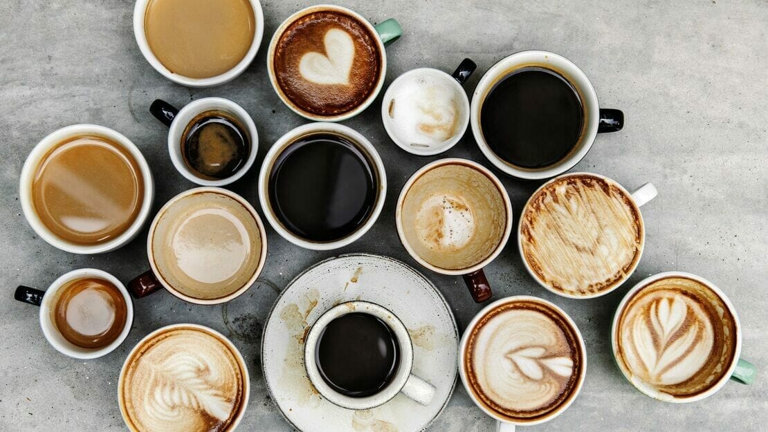 An assortment of coffee cups in various sizes and styles, arranged on a gray surface, some with latte art and some plain, ready to fuel your day at full speed.