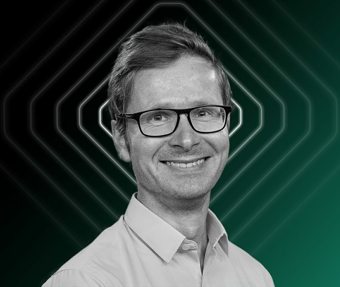 A man with glasses and a slight smile, wearing a light-colored shirt. The background features geometric patterns with a gradient from dark to light green, subtly reminiscent of the dynamic adaptability of Spryker on AWS.