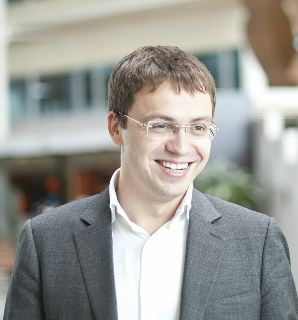 A man with short brown hair and glasses smiles while wearing a grey suit and white shirt, ready to excite experts with his confident demeanor. The background features an indoor setting with blurred details.