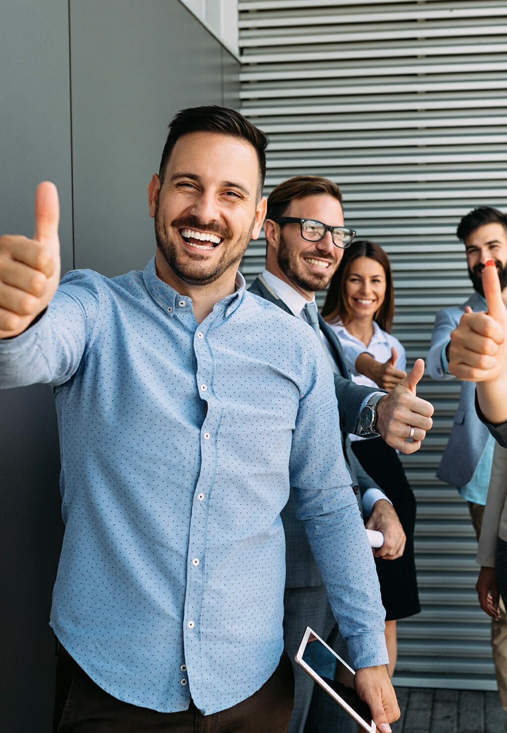 A group of six diverse professionals stand in line, smiling and giving thumbs up gestures in a modern, industrial-looking setting, embodying the spirit of emerce.