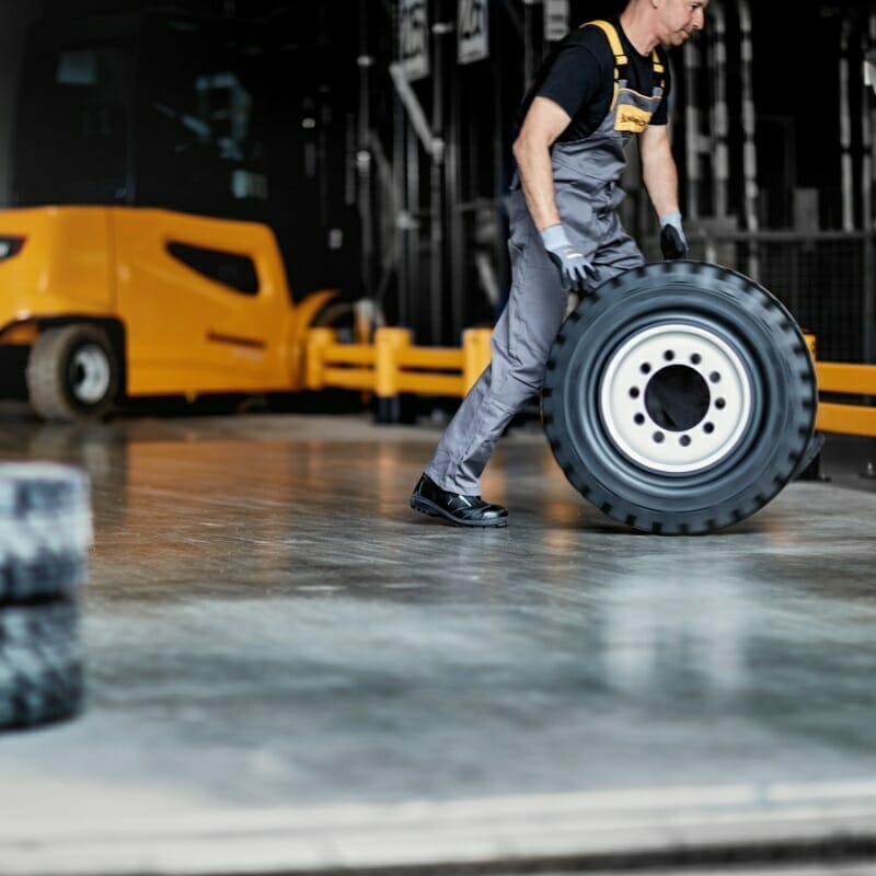 A person in work attire is rolling a large tire inside an industrial setting with a yellow forklift in the background, showcasing the efficiency of Spryker's logistics solutions.