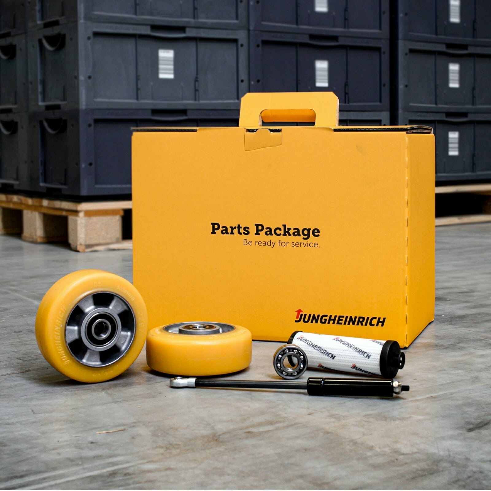 Yellow parts package with two wheels, a hydraulic cylinder featuring the Jungheinrich brand name, and various other components, all positioned in front of a stack of crates. Spryker innovation meets robust engineering.