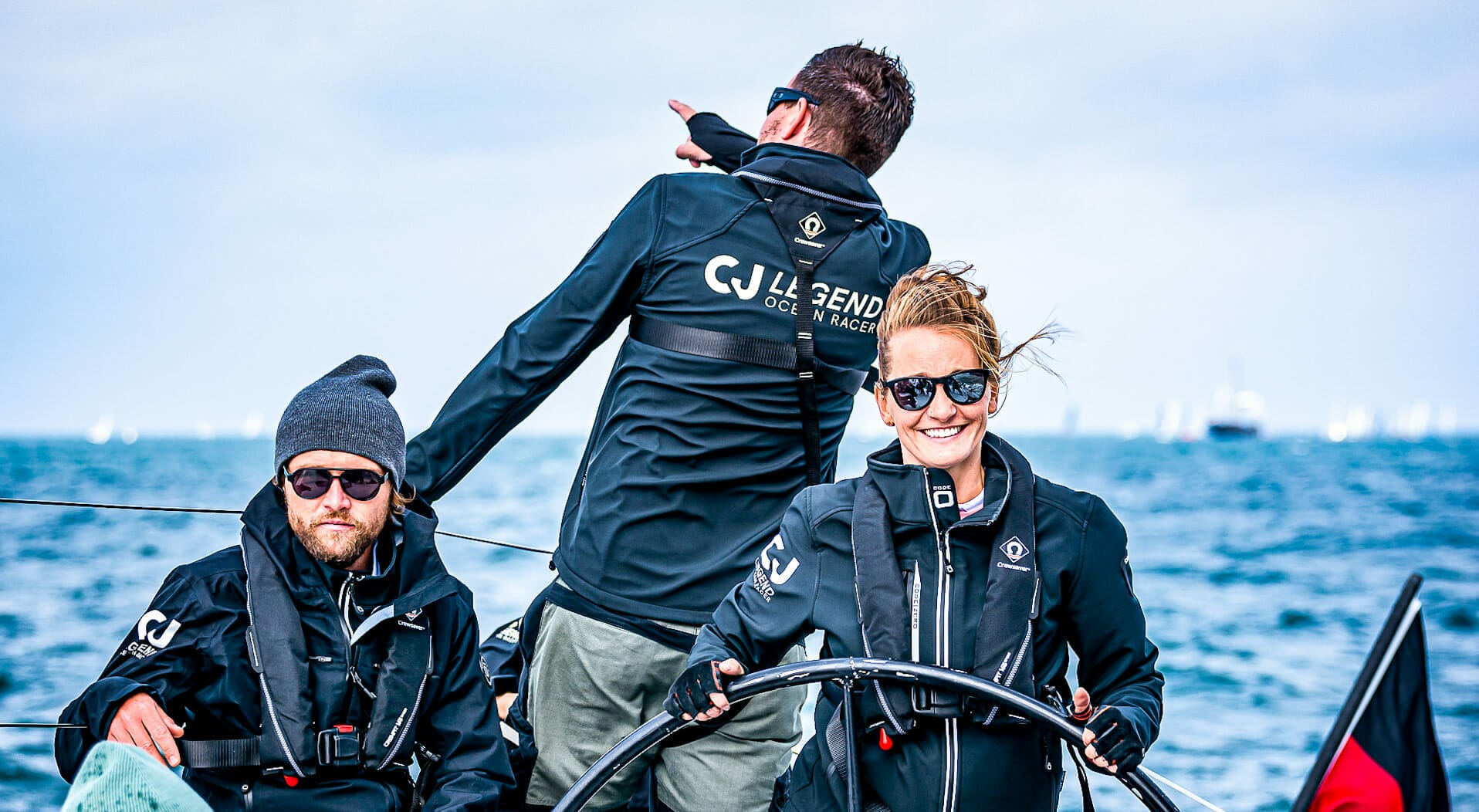 Three sailors on a boat wearing dark jackets are immersed in their sailing adventure. One is steering, another is seated looking ahead, and the third is standing and pointing forward. The ocean and other boats create a stunning backdrop for this voyage.