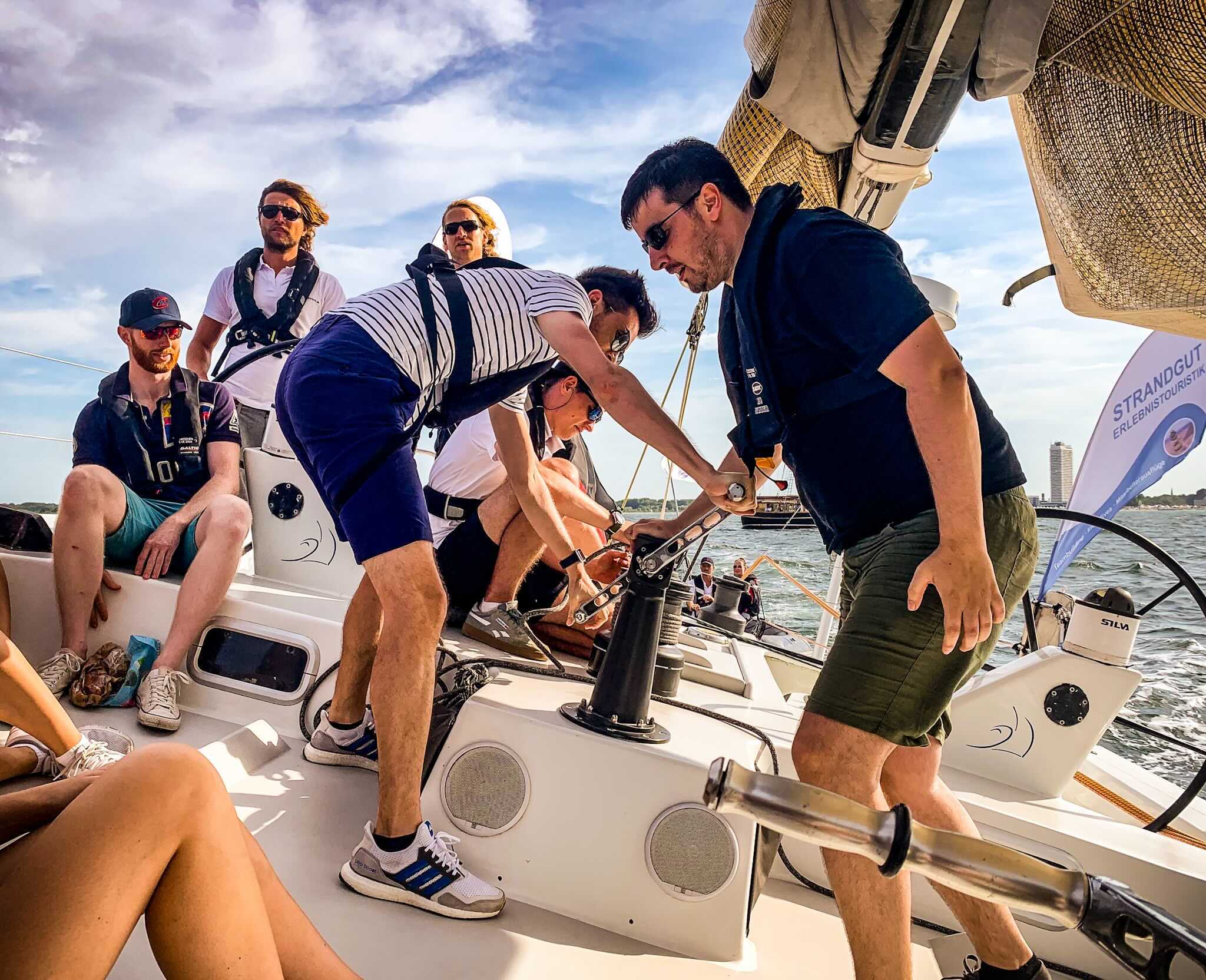 A group of people are engaged in sailing on a boat. Two men in the foreground are working with a winch, while the others watch or prepare to assist. The scene is set on a sunny day with a partly cloudy sky and the boat tilted to one side, indicating motion.