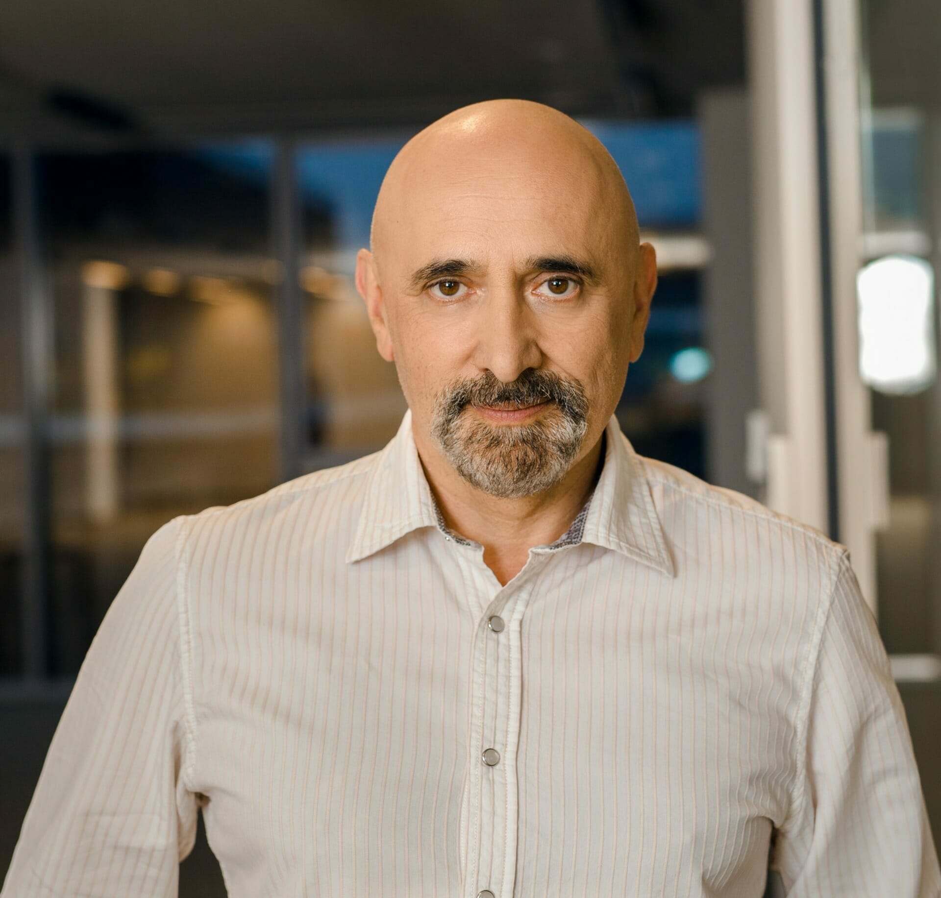 A bald man with a goatee, wearing a light-colored striped shirt, stands indoors in a well-lit environment, possibly discussing the latest trends in PaaS SaaS solutions.