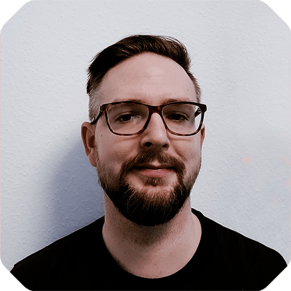 Man with short hair, glasses, and a beard standing against a plain white background, wearing a black t-shirt featuring the Spryker Marketplace logo, and looking at the camera.