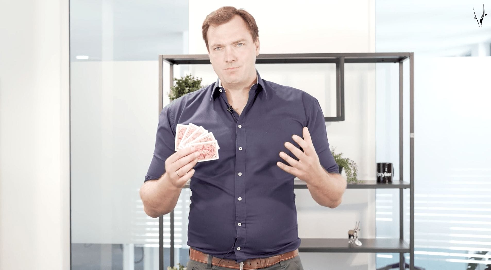 A person in a blue shirt holds a fan of playing cards in one hand and gestures like a magician with the other while standing in an e-commerce office space.