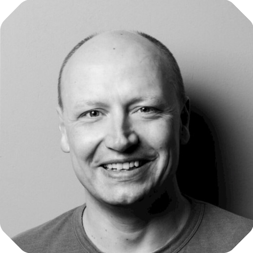 A black and white portrait of a smiling man with a bald head, wearing a casual shirt, set against the plain backdrop of a B2B suite.