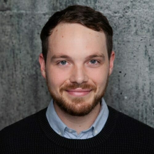 A man with short dark hair and a beard is wearing a light blue collared shirt under a black sweater, standing in front of a mottled gray concrete wall.