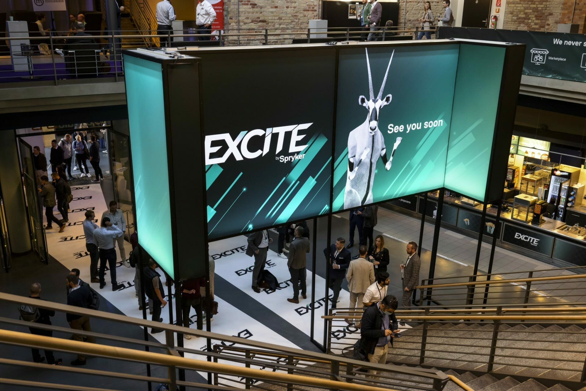 At Shoptalk Las Vegas, attendees stroll through an exhibit hall. A large display screen prominently features the brand "EXC!TE" with the slogan "See you soon" and an image of a goat.