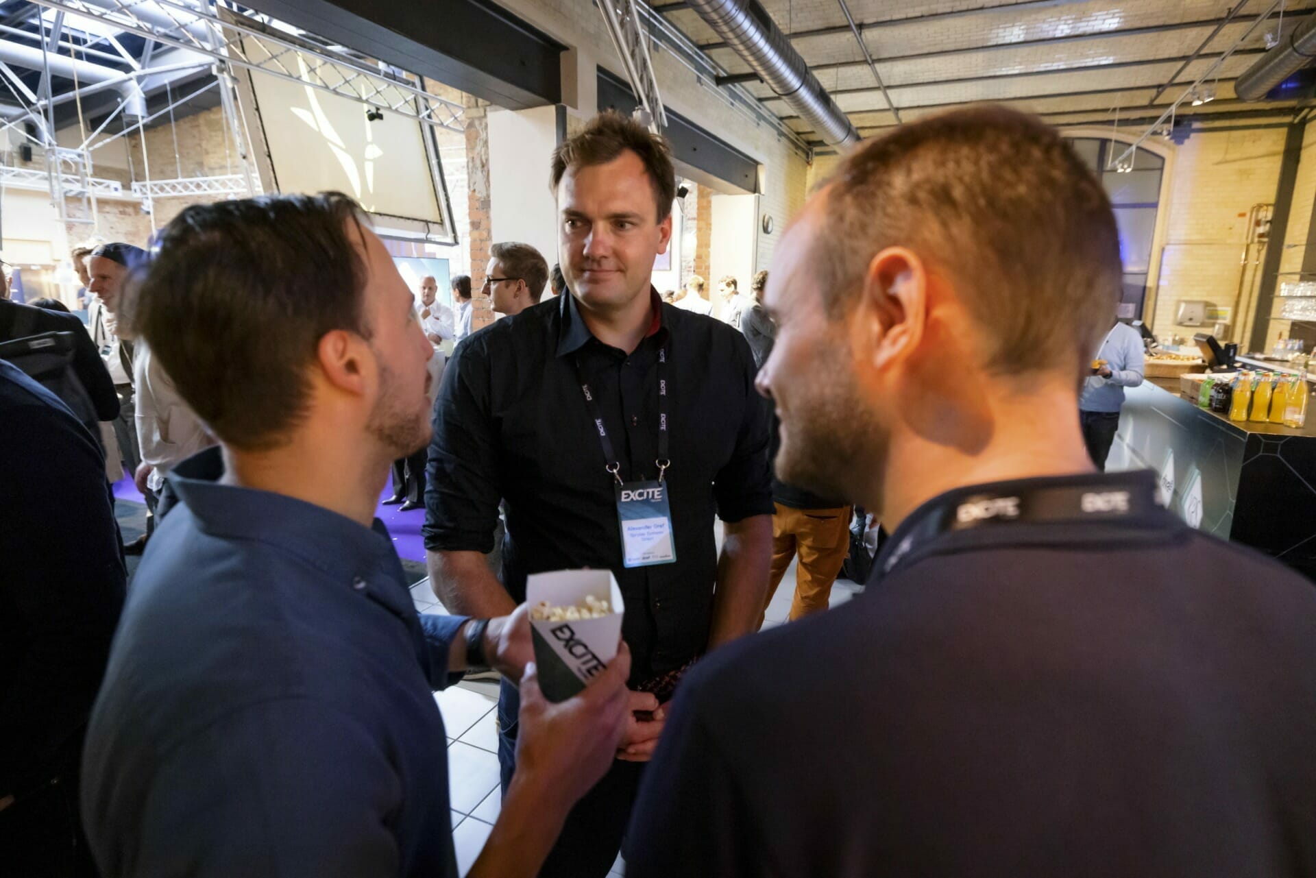 Three men are deep in shoptalk at an indoor event, with one man holding a container of popcorn. A banner and people are visible in the bustling background.