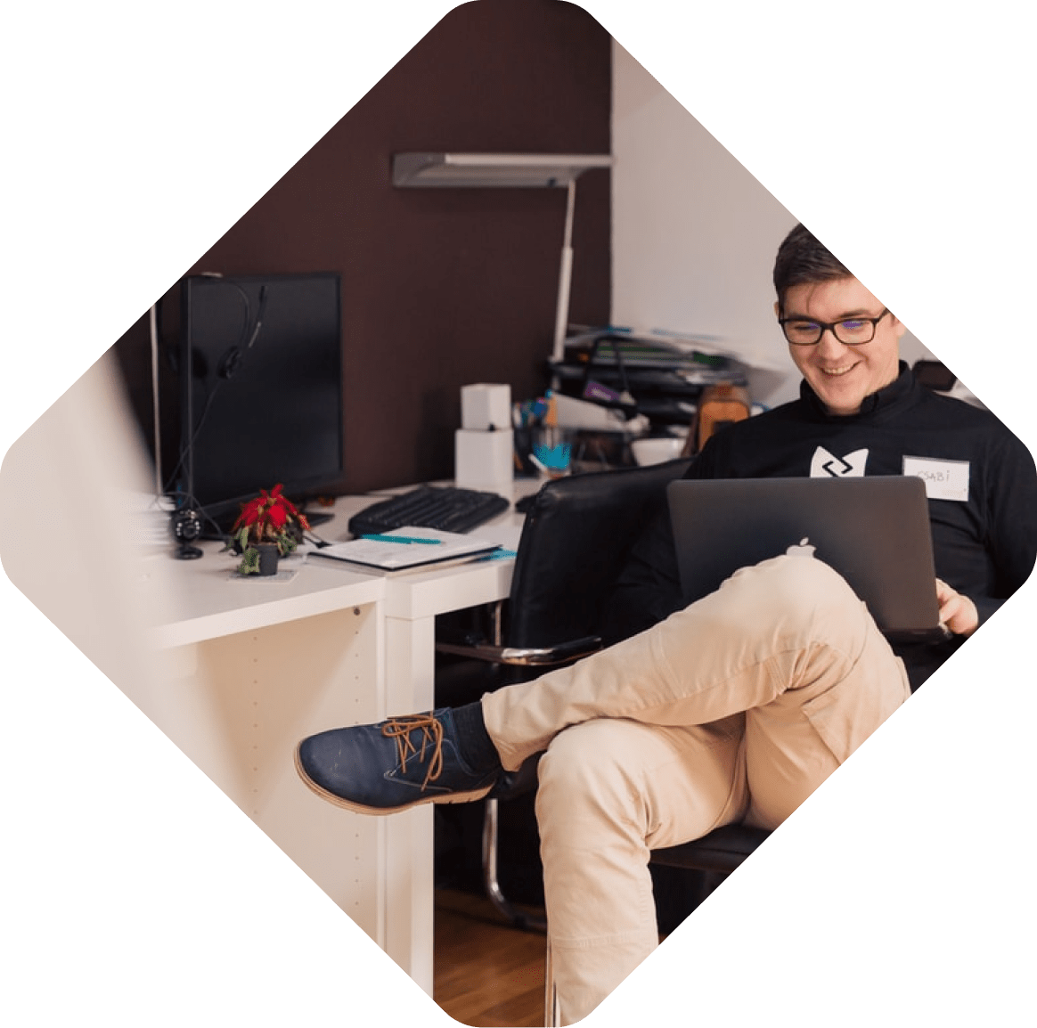 A person sitting on a chair, working on a laptop in their home office, surrounded by a desk, computer monitor, and various items nearby. With an eye focused on the screen, they're delving into the complexities of composable commerce.