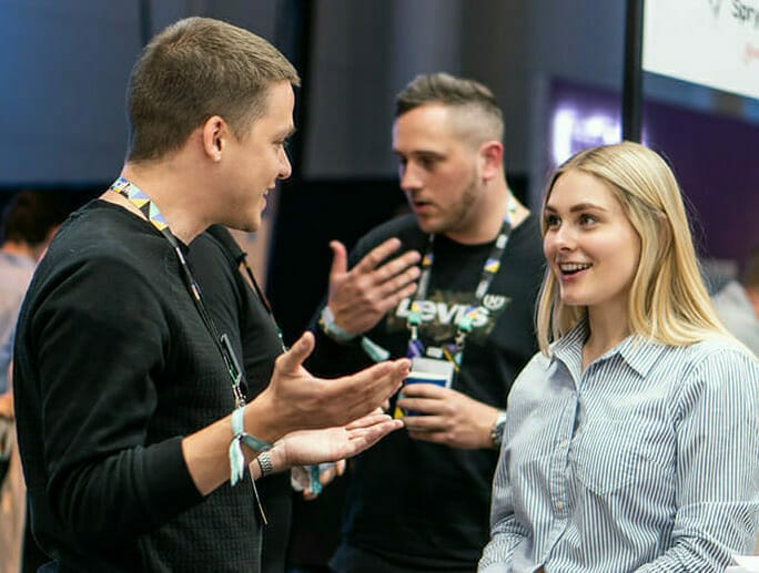 Two men and a woman engage in conversation at an indoor event. The woman smiles while one man gestures as he speaks, and the other man stands behind them talking, holding a drink with an NRF badge clipped to his lapel.