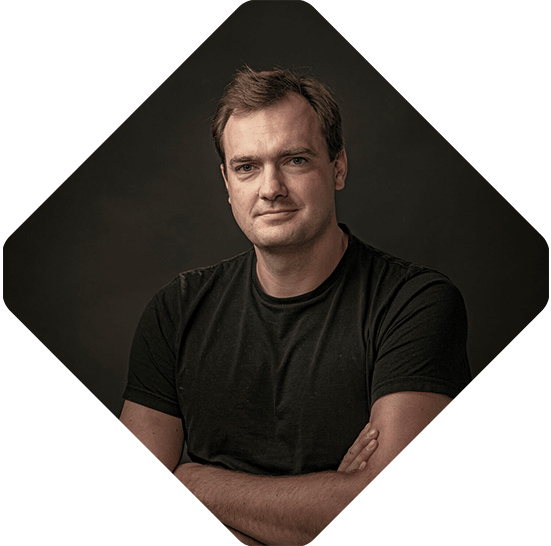 A man with short hair and a serious expression stands with his arms crossed, wearing a black t-shirt against a dark background, looking as if he's about to engage in intense commerce talks.