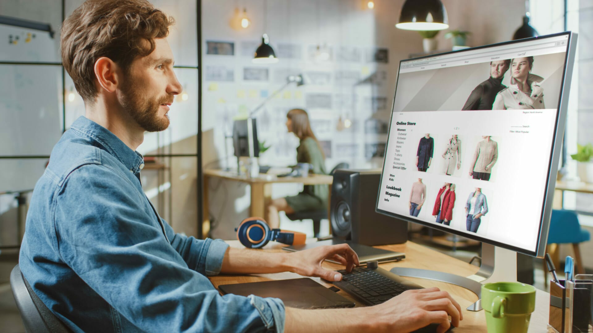 A person, the e-commerce champion, uses a desktop computer to browse an online clothing store in a well-lit office while another individual works in the background.